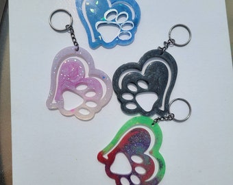 May include: Four heart-shaped keychains with a paw print design. The keychains are made of clear resin with different colors and glitter. The colors are blue, pink, black, and green.