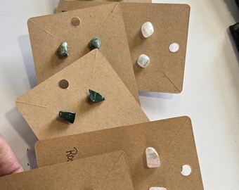 May include: A collection of stud earrings featuring various polished natural gemstones mounted on brown rectangular cardboard display cards. The stones include tiger's eye, green aventurine, white moonstone, dark green malachite, light pink rose quartz, and black obsidian.
