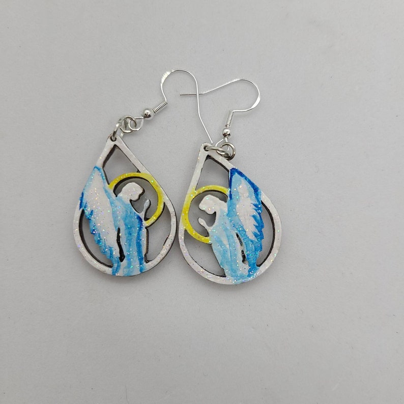 May include: A pair of silver dangle earrings with a teardrop-shaped design. Each earring features a white background with a blue and yellow angel silhouette.