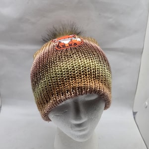 May include: A knitted beanie hat with a brown, green, and pink color scheme. The hat features a fox patch and a fluffy, dark brown pom-pom on top. The hat is displayed on a white mannequin head.