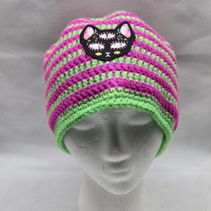 May include: A handmade crochet beanie in bright pink and green stripes. The hat features a black cat patch with pink ears and white details. The brim is green, and the hat is displayed on a white mannequin head.