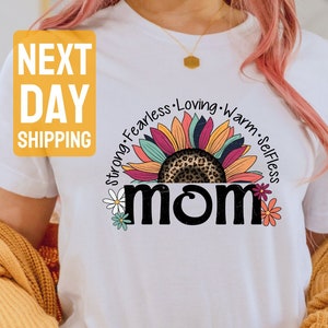 May include: White t-shirt with a colorful sunflower design and the word "MOM" in black text. The sunflower has a leopard print center and the words "Strong, Fearless, Loving, Warm, Selfless" are written around the sunflower.