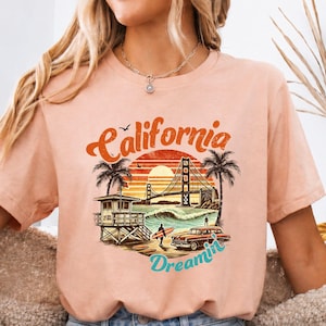 May include: Heather peach t-shirt featuring a California Dreamin' graphic. The design includes a sunset, palm trees, the Golden Gate Bridge, a lifeguard tower, and a vintage car. The shirt is a soft, comfortable material.