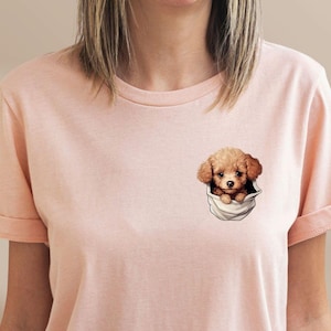 May include: Peach-colored t-shirt featuring a pocket design with a cute brown puppy peeking out. The shirt has short sleeves and a relaxed fit. The word "Peach" is in a circle in the upper left corner.