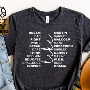 May include: A dark heather grey t-shirt with white text. The text reads "Dream, Lead, Fight, Write, Speak, Dare, Think, Reclaim, Educate, Challenge, Inspire" on the left side of the shirt. On the right side of the shirt, the text reads "Martin, Harriet, Malcolm, Maya, Frederick, Shirley, Garvey, Maxine, W.E.B., Rosa, Obama".