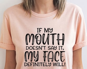 May include: Heather peach-colored t-shirt with the text "IF MY MOUTH DOESN'T SAY IT, MY FACE DEFINITELY WILL!" in black. The shirt has rolled-up sleeves and is made of a soft material. The shirt is a casual wear item.
