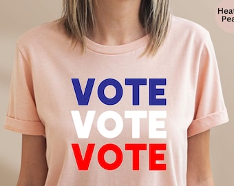May include: A heather peach short-sleeve t-shirt featuring the word VOTE printed three times in a stacked design. The top VOTE is in blue, the middle VOTE is in white, and the bottom VOTE is in red, creating a patriotic color scheme on the shirt.