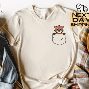 May include: A beige t-shirt with a cartoon image of a brown squirrel peeking out of a front pocket. The t-shirt has a crew neck and short sleeves.