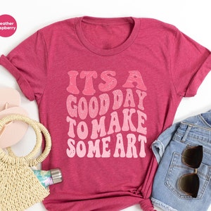 May include: A heather raspberry t-shirt with the text "It's a good day to make some art" in a pink and white gradient font.