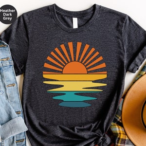 May include: Heather dark grey t-shirt featuring a retro sunset design. The graphic shows a vibrant orange sun with radiating lines above a layered reflection in shades of orange, yellow, and teal. The text "Heather Dark Grey" is in the upper left corner.
