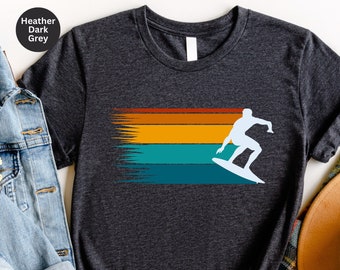 May include: Heather dark grey t-shirt featuring a retro design. The design includes a white silhouette of a surfer on a surfboard, set against a horizontal stripe of orange, yellow, teal, and blue. The shirt has a crew neck and short sleeves.