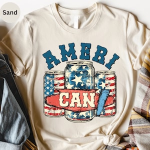 May include: Sand-colored t-shirt featuring a patriotic design. The graphic includes three beverage cans with American flag and star patterns, and the text "AMERI CAN" in blue and red. The shirt is made of a soft material.