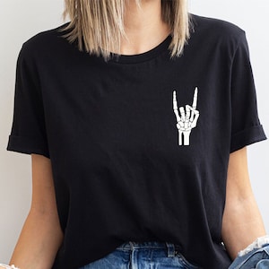May include: Black t-shirt featuring a small, white skeleton hand graphic making a rock-and-roll hand gesture. The shirt has short sleeves and a relaxed fit.  The image shows the shirt on a model, highlighting its casual style and the subtle yet eye-catching design. The graphic is simple and stylish, suitable for fans of rock music and alternative fashion.