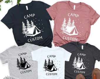 May include: Five t-shirts with a camping theme. The shirts are in different colours, including black, grey, white, and pink. Each shirt features a white silhouette of a tent and trees with the text "Camp Custom".