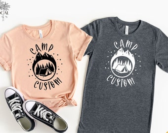 May include: Two t-shirts with a camping theme. The shirt on the left is heather peach with black text and graphics. The shirt on the right is heather dark grey with white text and graphics. Both shirts feature a tent, mountains, and trees inside a circle with the text "Camp Custom".