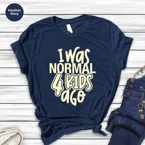 Może przedstawiać: Granatowa koszulka z napisem "I Was Normal 4 Kids Ago" w kremowym kolorze. Koszulka jest złożona, z dżinsami i butem widocznymi w tle. Koszulka wykonana jest z miękkiego materiału.