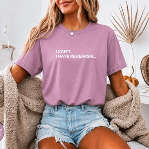 May include: Heather orchid colored t-shirt with the text "I can't. I have rehearsal." The shirt is paired with denim shorts and a beige cardigan. A decorative pumpkin and dried plant are in the background.