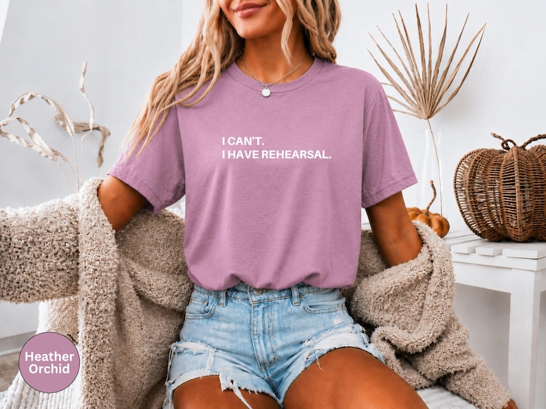 May include: Heather orchid colored t-shirt with the text "I can't. I have rehearsal." The shirt is paired with denim shorts and a beige cardigan. A decorative pumpkin and dried plant are in the background.