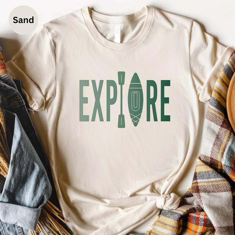 Puede incluir: Una camiseta color arena con la palabra "EXPLORE" en letras verdes. La letra "O" se reemplaza con un gr&aacute;fico de kayak y remo. La camiseta est&aacute; hecha de un material suave y es adecuada para uso casual.