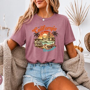 May include: Heather mauve t-shirt with a California graphic. The design features a sunset, palm trees, the Golden Gate Bridge, and the word "California" in orange and blue. The shirt is a short-sleeved crew neck.