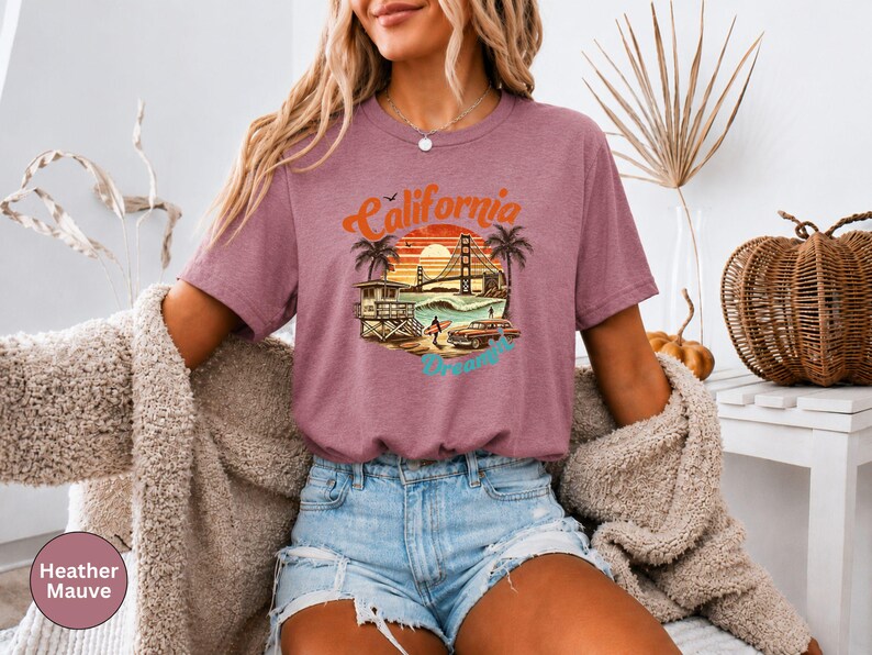 May include: Heather mauve t-shirt with a California graphic. The design features a sunset, palm trees, the Golden Gate Bridge, and the word "California" in orange and blue. The shirt is a short-sleeved crew neck.