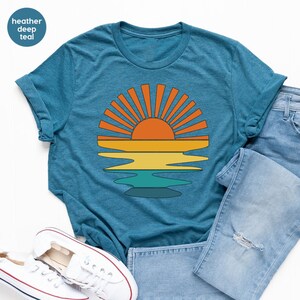 May include: Heather deep teal t-shirt featuring a retro sunset graphic. The design includes an orange sun with yellow, orange, and teal water reflections. The shirt is paired with blue jeans and white sneakers.