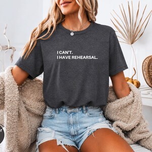 May include: Heather dark grey t-shirt with the text "I can't. I have rehearsal." in white. The shirt is a crew neck and has short sleeves. The model is wearing blue jean shorts and a beige cardigan.