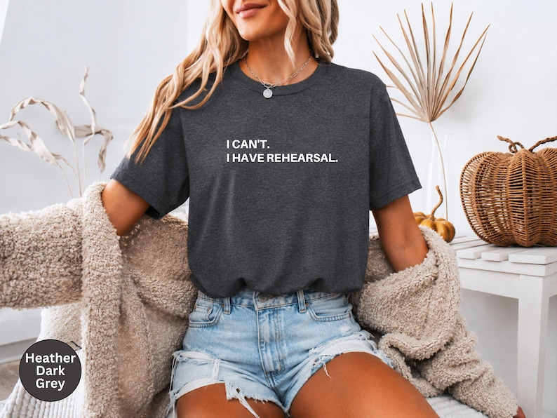 May include: Heather dark grey t-shirt with the text "I can't. I have rehearsal." in white. The shirt is a crew neck and has short sleeves. The model is wearing blue jean shorts and a beige cardigan.