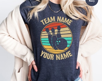 May include: A heather navy blue t-shirt with a retro-style graphic of bowling pins and a bowling ball. The graphic is surrounded by a circle with the text "TEAM NAME" above and "YOUR NAME" below.