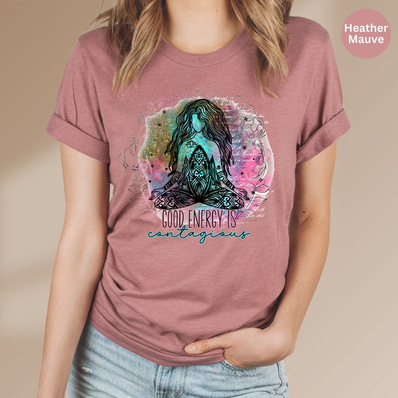 Peut inclure: T-shirt mauve chin&eacute; avec des manches retrouss&eacute;es. Le t-shirt pr&eacute;sente un graphisme d'une figure m&eacute;ditant avec de longs cheveux, entour&eacute;e d'un motif aquarelle. Le texte "GOOD ENERGY IS contagious" est en dessous.