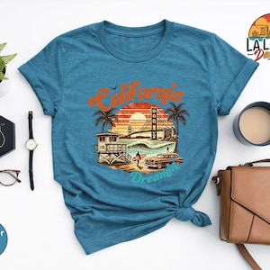 May include: A teal t-shirt featuring a California-themed graphic. The design includes the Golden Gate Bridge, palm trees, a lifeguard tower, a vintage car, and the text "California Dreamin'". The shirt is styled with accessories.