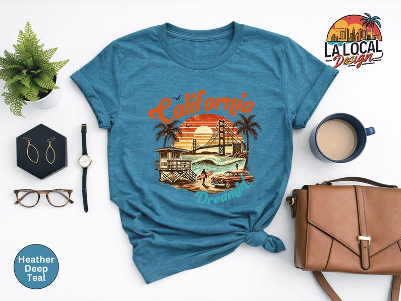 May include: A teal t-shirt featuring a California-themed graphic. The design includes the Golden Gate Bridge, palm trees, a lifeguard tower, a vintage car, and the text "California Dreamin'". The shirt is styled with accessories.