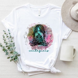 Peut inclure: T-shirt blanc avec une illustration color&eacute;e d'une figure m&eacute;ditative aux longs cheveux, entour&eacute;e d'&eacute;l&eacute;ments floraux. Le texte "GOOD ENERGY IS contagious" est imprim&eacute; en dessous. Un chapeau et une tasse blanche sont visibles.