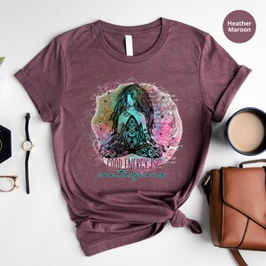 Peut inclure: T-shirt bordeaux chin&eacute; avec un motif color&eacute; d'une figure m&eacute;ditant avec de longs cheveux, entour&eacute;e d'&eacute;l&eacute;ments floraux. Le texte "GOOD ENERGY IS contagious" est en dessous. Une montre, des lunettes et un sac &agrave; main sont en arri&egrave;re-plan.