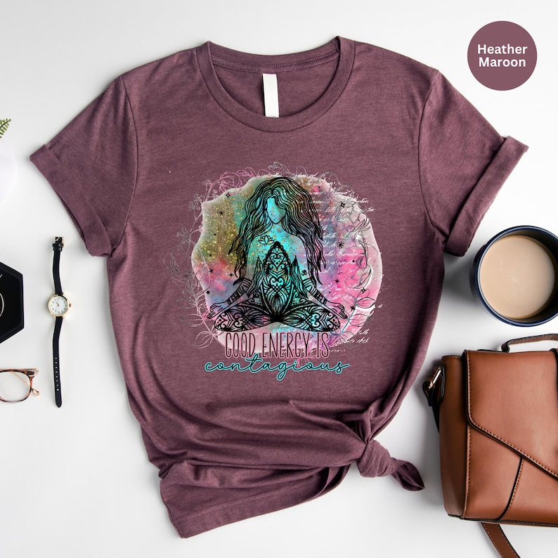 Peut inclure: T-shirt bordeaux chin&eacute; avec un motif color&eacute; d'une figure m&eacute;ditant avec de longs cheveux, entour&eacute;e d'&eacute;l&eacute;ments floraux. Le texte "GOOD ENERGY IS contagious" est en dessous. Une montre, des lunettes et un sac &agrave; main sont en arri&egrave;re-plan.