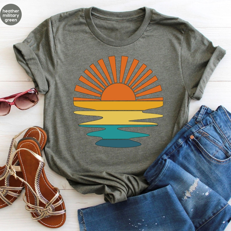 May include: Heather military green t-shirt featuring a retro sunset design. The graphic shows a vibrant orange sun with radiating lines above a colorful horizon of yellow, orange, and teal. The shirt is paired with blue jeans and sandals.