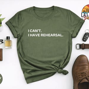May include: Olive green t-shirt with the text "I CAN'T. I HAVE REHEARSAL." in white. The shirt is styled with a brown belt, shoes, wallet, glasses, and a small potted plant. The shirt is a military green color.