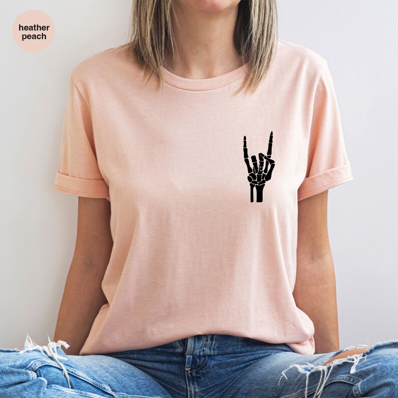 May include: A peach-colored t-shirt with rolled-up sleeves. A black skeleton hand graphic is printed on the front, displaying the rock and roll hand gesture. The upper left corner of the shirt has the text "heather peach".