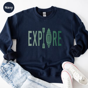 Puede incluir: Sudadera azul marino con la palabra "EXPLORE" en letras verdes. La "O" se sustituye por un dibujo de kayak y remo. La sudadera se muestra con vaqueros azul claro y zapatillas blancas.