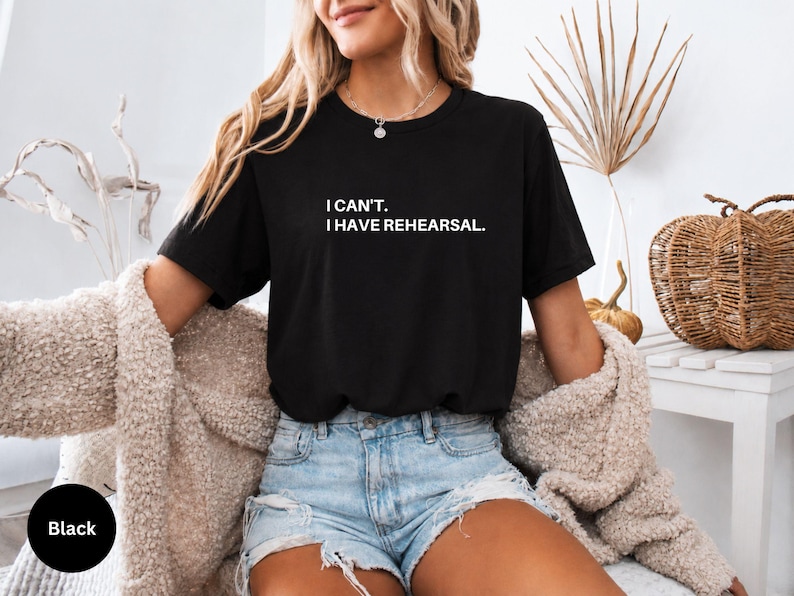 May include: Black t-shirt with the text "I can't. I have rehearsal." in white. The shirt is worn with denim shorts and a beige cardigan. A black circle with the word "Black" is in the lower left corner.