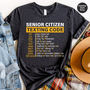 May include: A dark grey t-shirt with the text "Senior Citizen Texting Code" printed in yellow. The shirt lists common texting abbreviations and their meanings for senior citizens.