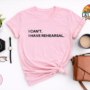 May include: Light pink t-shirt with the text "I CAN'T. I HAVE REHEARSAL." in black. The shirt is styled with accessories including a watch, earrings, glasses, a purse, and a coffee cup. The LA Local Design logo is in the upper right corner.
