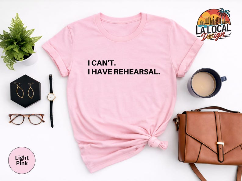May include: Light pink t-shirt with the text "I CAN'T. I HAVE REHEARSAL." in black. The shirt is styled with accessories including a watch, earrings, glasses, a purse, and a coffee cup. The LA Local Design logo is in the upper right corner.