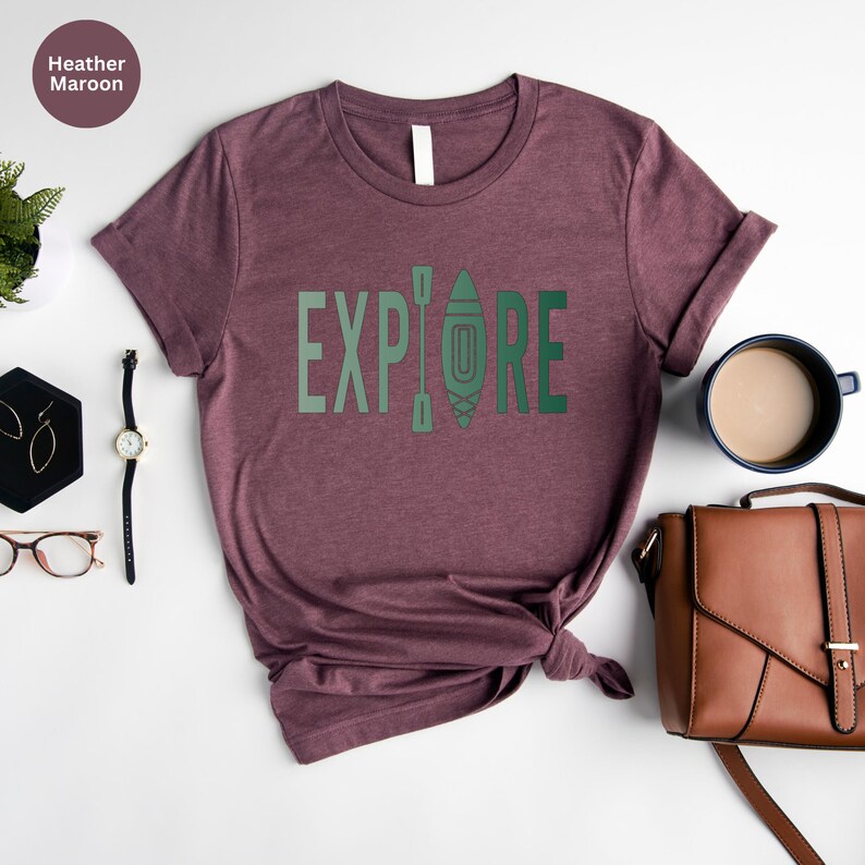 Puede incluir: Camiseta color borgo&ntilde;a con la palabra "EXPLORE" en verde y un gr&aacute;fico de kayak. La camiseta se muestra con accesorios que incluyen un reloj, pendientes y un bolso marr&oacute;n. Tambi&eacute;n se ve una taza de caf&eacute;.