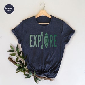 Puede incluir: Camiseta azul marino jaspeado con la palabra "EXPLORE" en letras verdes. La letra "O" est&aacute; reemplazada por un gr&aacute;fico de kayak y remos. La camiseta est&aacute; colgada en una percha de madera.