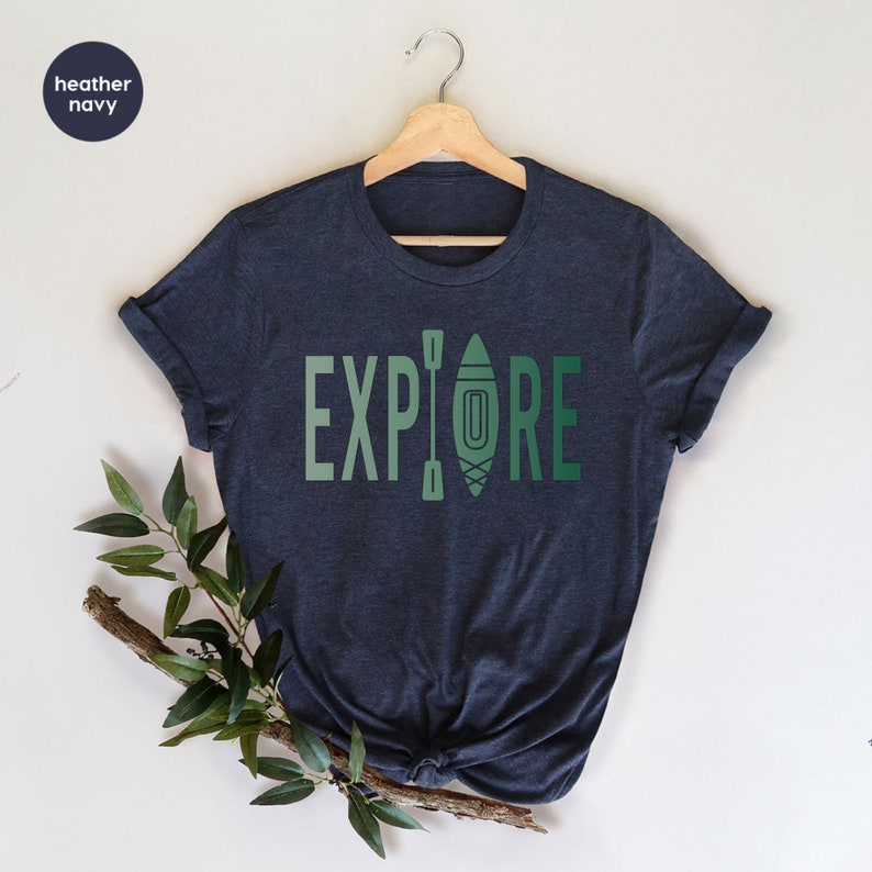 Puede incluir: Camiseta azul marino jaspeado con la palabra "EXPLORE" en letras verdes. La letra "O" est&aacute; reemplazada por un gr&aacute;fico de kayak y remos. La camiseta est&aacute; colgada en una percha de madera.