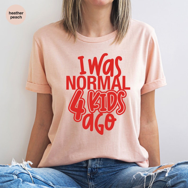 Może przedstawiać: Brzoskwiniowa koszulka z czerwonym napisem "I Was NORMAL 4 KIDS Ago". Koszulka ma podwinięte rękawy i jest wykonana z miękkiego materiału. Jest to element odzieży codziennej.