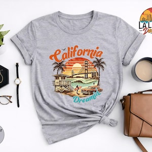 May include: Heather light grey t-shirt featuring a colorful graphic of the Golden Gate Bridge, palm trees, and a vintage car with the text "California Dreamin'". The shirt is styled with rolled-up sleeves and a knot at the waist.