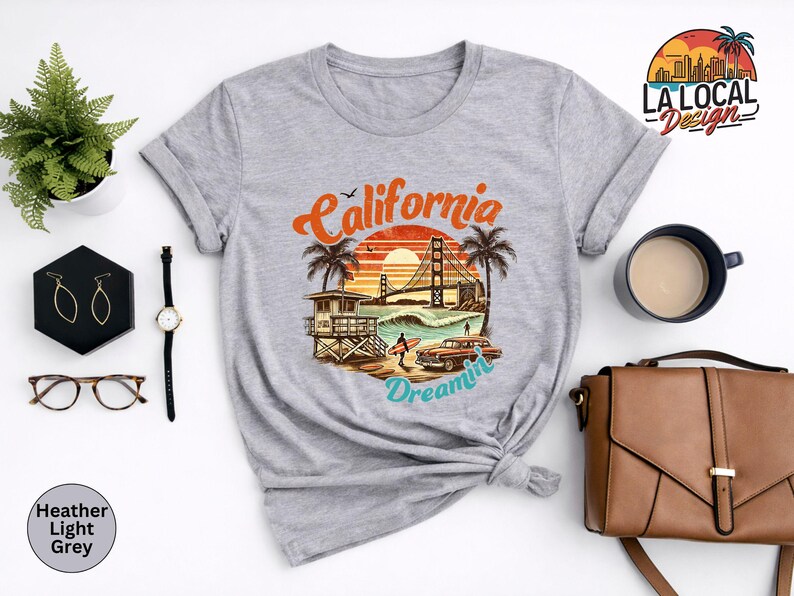 May include: Heather light grey t-shirt featuring a colorful graphic of the Golden Gate Bridge, palm trees, and a vintage car with the text "California Dreamin'". The shirt is styled with rolled-up sleeves and a knot at the waist.