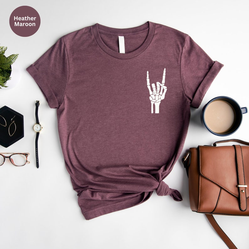May include: Heather maroon t-shirt with a white skeleton hand graphic making a rock and roll gesture. The shirt is displayed on a white surface with accessories including a watch, earrings, and a brown purse.
